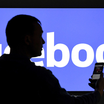 Muž s mobilním telefonem před logem společnosti Facebook - ilustrační foto.